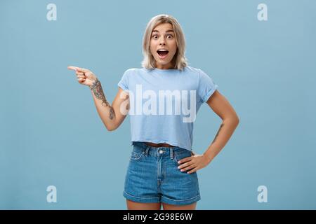 Studio shot of amazed impressed good-looking sportswoman in trendy t-shirt with tattoos opening mouth with enthusiasm from surprise holding hand on Stock Photo
