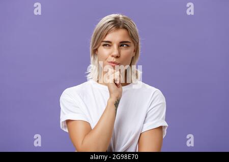 Waist-up shot of smart creative young ambitious woman with blond haircut and tanned skin squinting and smirking gazing left while holding hand on chin Stock Photo