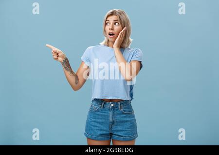 Studio shot of nervous shocked good-looking young woman with fair hair and tatooed arm gasping pointing and looking left with worry holding hand on Stock Photo