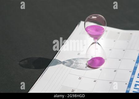 Hour glass with pink sand flowing and calendar on black tabletop background. Copy space. Stock Photo