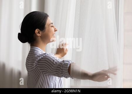 Happy dreamy beautiful Indian woman opening curtains, starting new day Stock Photo