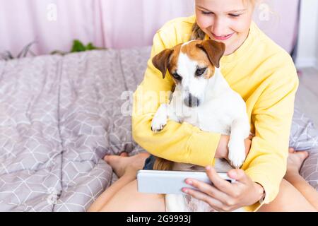 A smiling girl with a mobile phone in her hand, together with her dog Jack Russell Terrier, communicates on social media or via Zoom, makes a selfie Stock Photo
