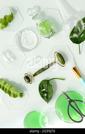 Self made moisturizer, green jade face roller and scissors with pieces of ice. Exotic monstera leaves and water drops on white background. Facial mass Stock Photo