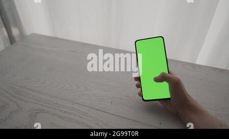 Man showing and using smarphone with green screen while sitting behind white oak table, wide photo Stock Photo