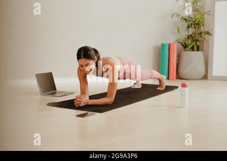 Woman in plank position looking at her mobile phone on floor. Woman doing pilates workout at home watching video on mobile phone. Stock Photo