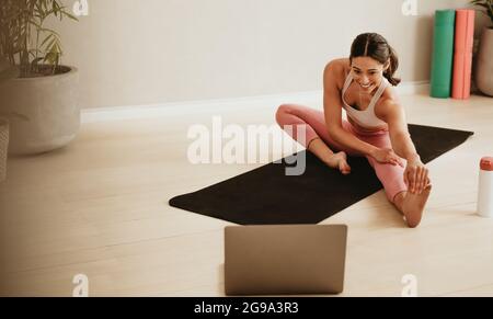 Female fitness trainer teaching leg stretching exercises online on laptop. Woman working out in front of her laptop in fitness studio. Stock Photo
