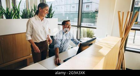 Two business professionals smiling and looking at computer. Mature woman standing next to young man working on computer. Stock Photo