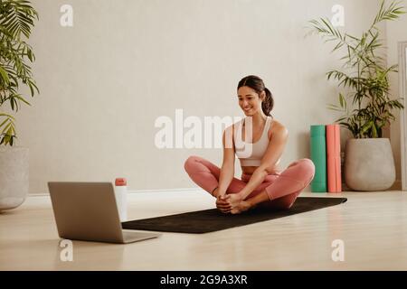 Woman sitting on yoga man and doing stretching workout while walking online video on laptop. Sporty woman doing exercises at home while watching onlin Stock Photo