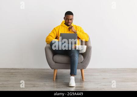 Black Man Using Laptop Sitting In Armchair Near Gray Wall Stock Photo