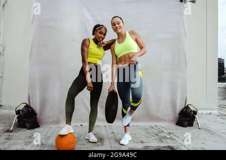 Sportswoman with foot on basketball standing with her friend in holding weight plate and looking at camera on rooftop. Females in sports clothing posi Stock Photo