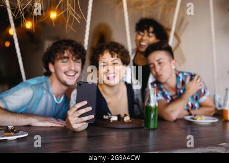 Group of friends taking a selfie together in a restaurant. Four young queer people smiling cheerfully while capturing the moment. Friends sitting and Stock Photo