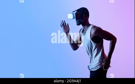 Virtual sports. Young black guy jogging while wearing VR headset in neon light, panorama with free space Stock Photo