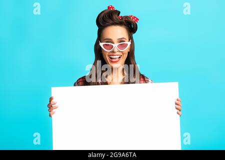 Shocked pinup woman in retro wear presenting something on white blank poster over blue studio background, mockup Stock Photo