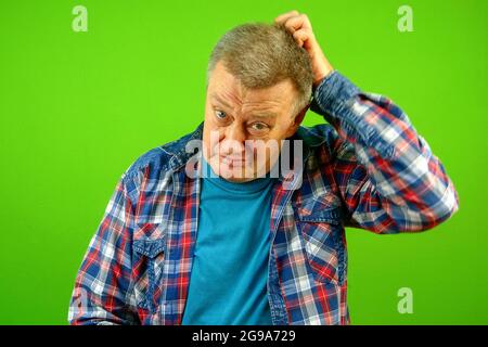 Confused puzzled, bewildered senior caucasian man in checkered shirt expression and scratching his head. He doubts. Green background. Close-up. Indoor Stock Photo