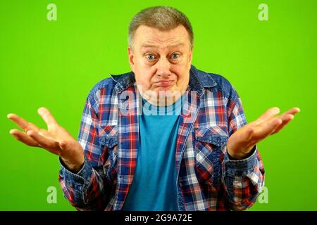 Confused puzzled, bewildered senior caucasian man in checkered shirt shrugging shoulders uncertain expression and spread arms. He doubts. Green backgr Stock Photo
