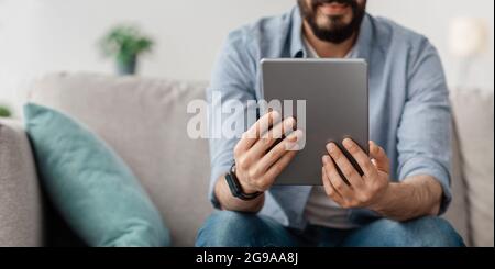 Unrecognizable man sitting on sofa, using nice mobile app on digital tablet, panorama, copy space, crop Stock Photo