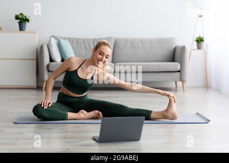Young blonde sports woman in sportswear doing workout on floor at home and watching fitness videos in laptop Stock Photo