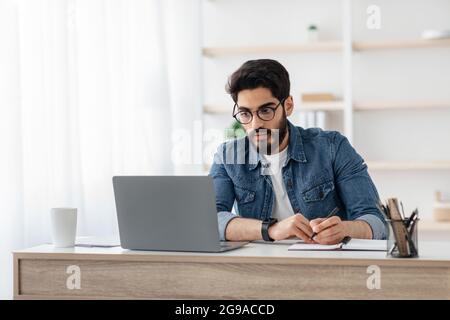Concentrated arab man taking notes and looking at laptop, writing project ideas in notebook sitting at workplace Stock Photo