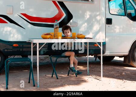 small boy in the shade of a tree in front of his camper sitting in a camping chair ready to eat the food on the table. Stock Photo