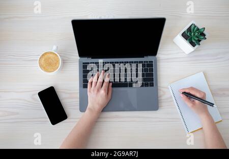 Office workspace. Unrecognizable businesswoman working at laptop and writing notes in notebook, top view Stock Photo