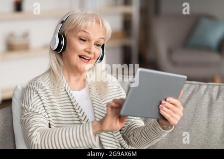 Smiling old lady in headset using tablet sitting on sofa Stock Photo