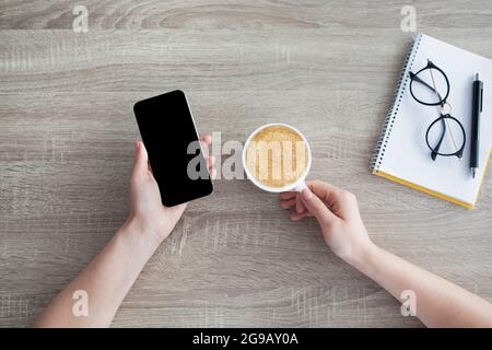 Modern lifestyle. Woman holsing smartphone with blank black screen and drinking coffee at workplace, top view Stock Photo