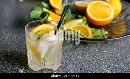 Lemon lemonade with mint and ice in a glass with black plastic tubes on a background of fruit. Cooling summer drink. horizontal close up Stock Photo