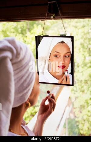 Beautiful young woman applying red lipstick while standing at the window and looking at mirror. Stock Photo