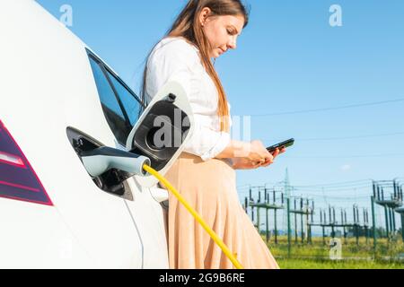 Woman plugging in the charger into her white electric car. Using a smartphone while waiting Stock Photo