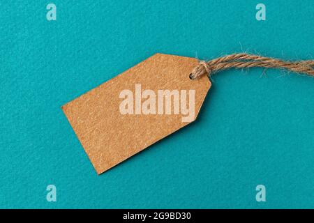Blank cardboard tag on a flax twine over turquoise background. Brown paper tag with jute rope against textured teal surface. Recyclable gift rope tag. Stock Photo
