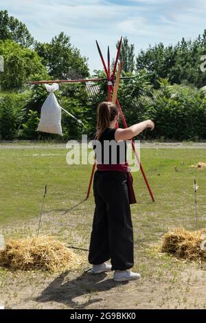 A young girl in a black suit with a red belt shoots from a vintage bow at the target from a bag with a straw. Cosplay festival in nature. Girl warrior Stock Photo