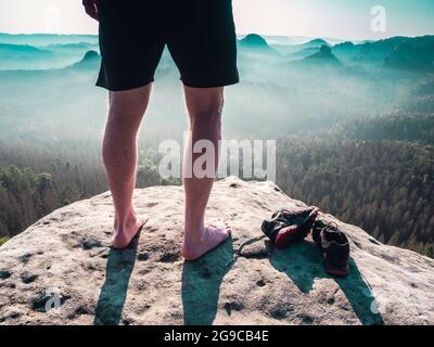 Barefoot slender legs with hairy calves of a male runner standing next to removed sweaty running shoes on a rocky edge above a long deep vally in natu Stock Photo