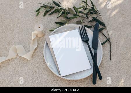 Festive wedding summer table setting. Black cutlery, olive branch, silk ribbon on dotted white dinner plate. Beige concrete background. Blank greeting Stock Photo