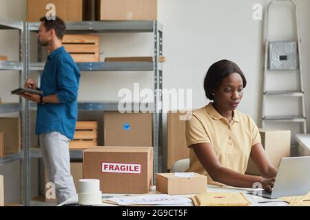 Portrait of young African-American woman using laptop in warehouse while managing shipping service, copy space Stock Photo