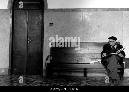 PARMA, ITALY - Mar 09, 2007: A grayscale shot of an elderly man sitting on a bench reading a book indoors in Parma, Italy Stock Photo