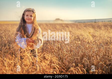 Young girl with traditional Bulgarian folklore costume at the agricultural wheat field during harvest time with industrial combine machine Stock Photo