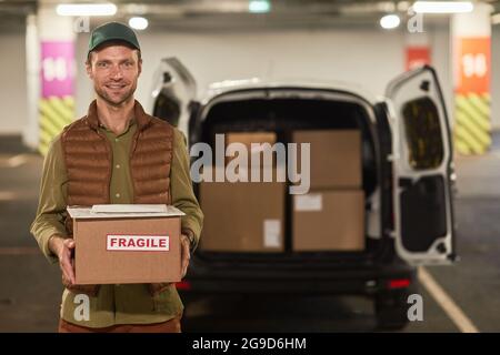 Waist up portrait of male delivery worker holding boxes and smiling at camera while standing by van outdoors, copy space Stock Photo