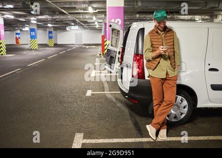 Full length portrait of male delivery worker standing by van at parking lot and using smartphone, copy space Stock Photo