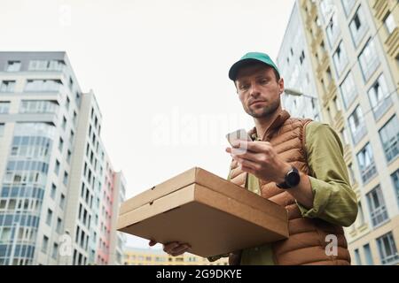 Low angle portrait of delivery man using smartphone and holding pizza while standing in urban setting, copy space Stock Photo