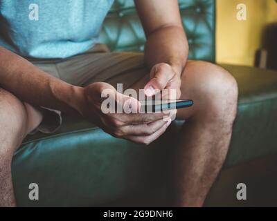 Hand man using smartphone. A young person, man in casual grey shirt and shorts sitting and interacting with technology at home. Relaxation businessman Stock Photo