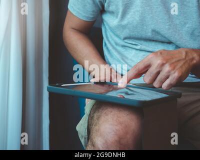 Hand man using digital tablet. A young person, man in casual grey shirt and shorts sitting and interacting with technology at home. Relaxation busines Stock Photo