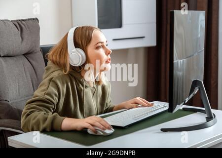 Teenager girl cybersportman gamer play online computer game at home alone in leisure time. Young blonde woman in headphones enthusiastically play pc Stock Photo