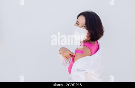 Vaccinations concept. Vaccinated Asian woman wearing medical face mask, pink sleeveless pointing at pink bandage plaster on her shoulder after vaccina Stock Photo