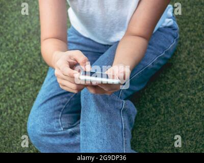 Mobile phone in hands of woman wearing white sleeveless shirt and blue jeans sitting with relax on green artificial grass background with copy space. Stock Photo