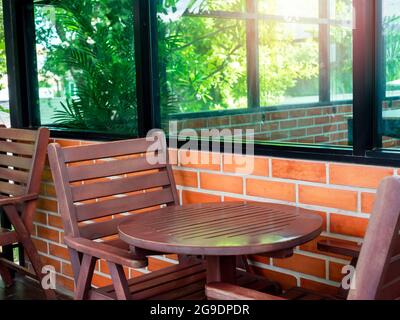 Empty red cozy wooden chairs and round wood coffee table near the glass window and brick wall with green garden in the cafe, summertime. Stock Photo
