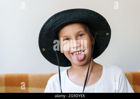 Portrait of charming caucasian boy in white t-shirt and black panama hat, in morning sunlight in the living room at home, sits on the couch, waiting for his parents. Childhood concept. School age boy Stock Photo