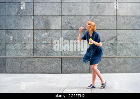 Carefree redhead woman celebrating blowing soap bubbles in town against a grey wall of a commercial building on a breezy day with copyspace Stock Photo