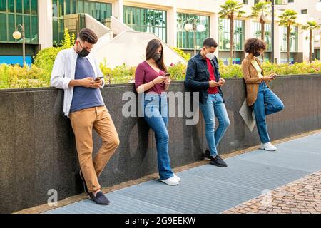 Line of friends ignoring each other to text on their mobile phones as they relax in an urban street leaning against a wall wearing protective face mas Stock Photo