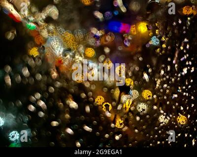 Shot through the glass in the rain with street lights and car headlights in the background. Blurred bokeh glitter of light background Stock Photo