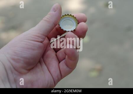 A hand holds the stopper of a soft drink against a blurry background. The left hand of an adult male holding a round metal bottle cap. Stock Photo